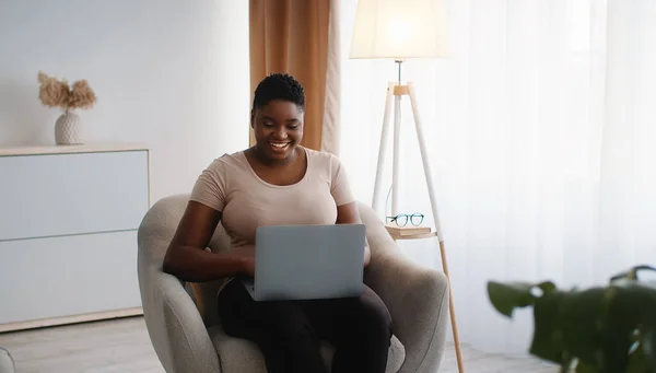 Молодая пухленькая африканская американка, работающая на ноутбуке, общающаяся с клиентами или партнерами, работающая на дому — стоковое фото