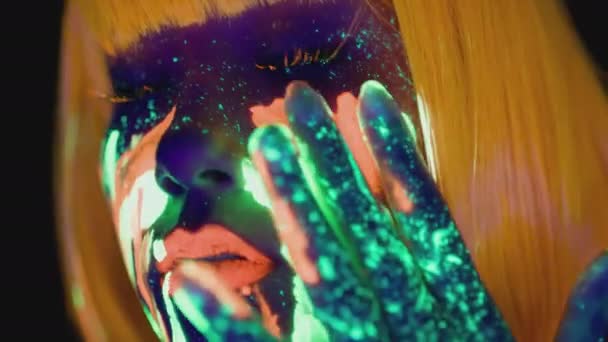 Lysande kroppskonst. Ung dam med neon makeup och orange peruk målning glödande färg på ansiktet, närbild porträtt — Stockvideo