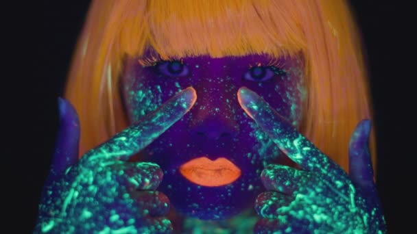 Neon body art. close-up van jonge vrouw dragen pruik smeren oranje verf op haar wangen, dansen in ultraviolet licht — Stockvideo