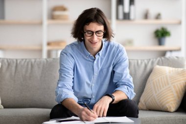 Not defteri olan pozitif bir erkek öğrenci evde oturuyor ve uzaktan çalışıyor, ödev yapıyor, boş yer var.