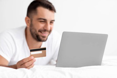 Beyaz tişörtlü mutlu beyaz beyaz erkek internetten alışveriş yapar, bilgisayardan sipariş verir ve kredi kartını gösterir.