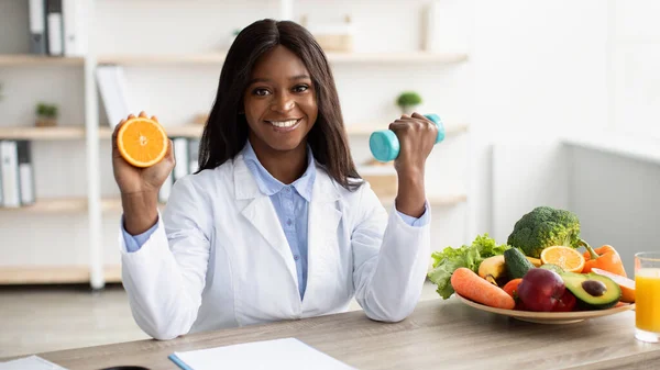 Felice donna afro-americana dietologa tenendo metà arancione e manubri in mano, raccomandando uno stile di vita sano — Foto Stock