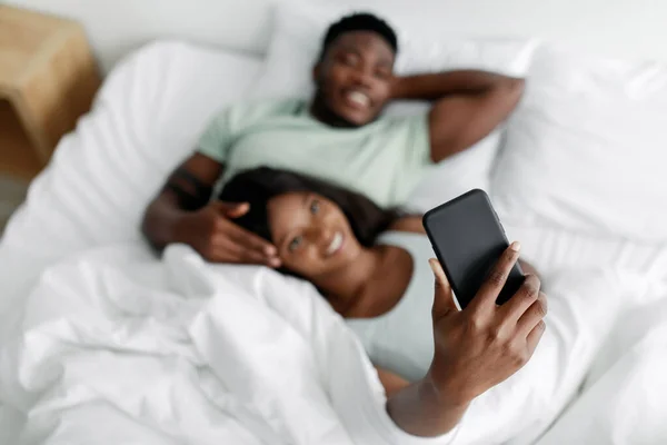Mosolygó fiatal afro-amerikai férj és feleség pihen az ágyon, feleség csinál szelfi a szociális média a hálószobában, elmosódott — Stock Fotó