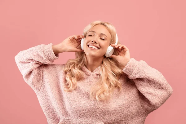 Klidná mladá žena poslouchající hudbu se zavřenýma očima, nošení a dotýkání sluchátek na růžovém pozadí studia — Stock fotografie