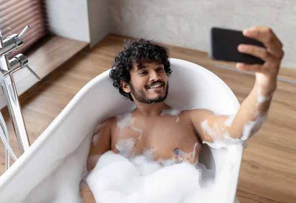 Hombre indio guapo acostado en la bañera con espuma y tomando selfie en el teléfono inteligente, descansando en el baño en casa — Foto de Stock