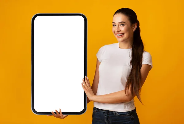 Mulher feliz segurando grande smartphone com tela vazia, fundo amarelo — Fotografia de Stock