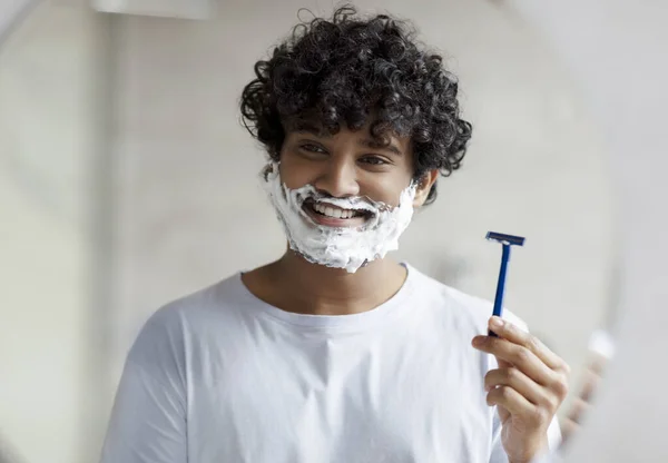 Fröhlicher indischer Mann mit einem Gesicht, das mit Rasierschaum bedeckt ist und einen Sicherheitsrasierer hält, während er im Badezimmer auf den Spiegel schaut — Stockfoto