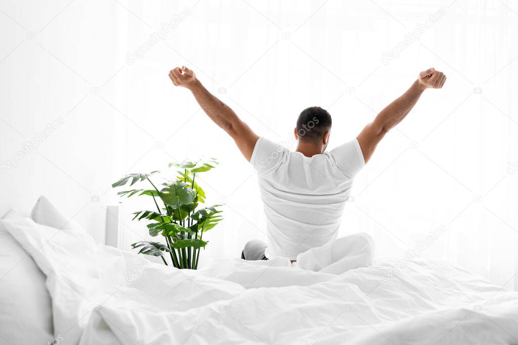 Satisfied european millennial guy wakes up after rest, stretching body on bed in bedroom interior, sun flare