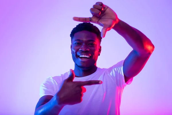 Χαρούμενος νεαρός μαύρος άντρας που φτιάχνει κορνίζα με δάχτυλα, κοιτάζει την κάμερα και χαμογελά για φωτογραφία στο νέον φως — Φωτογραφία Αρχείου