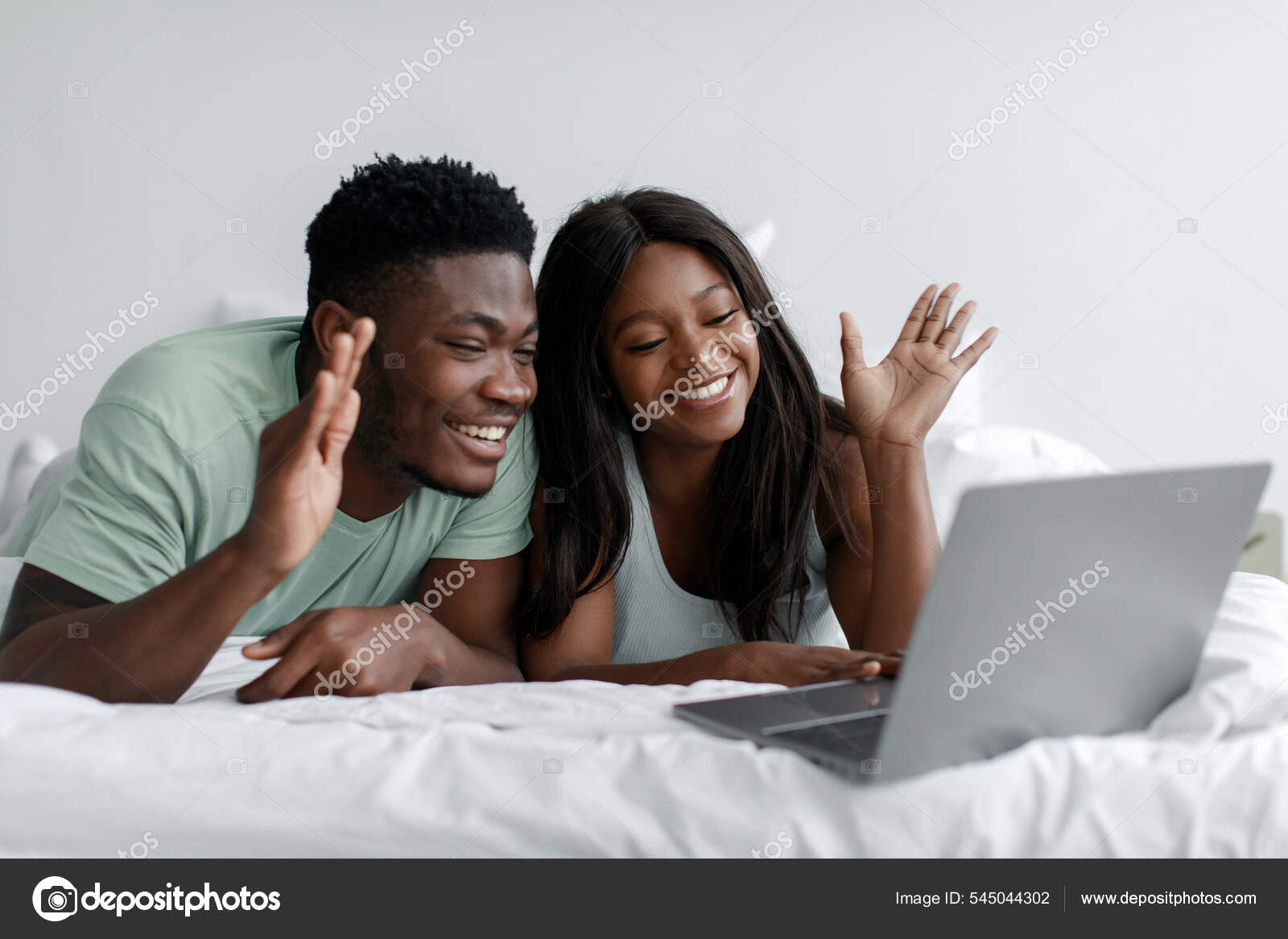 Cheerful young african american wife and husband lie on bed waving hand at  computer webcam in bedroom Stock Photo by ©Milkos 545044302