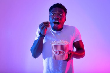 Heyecanlı genç zenci adamın portresi kovadan patlamış mısır yiyor, sinemada veya televizyonda film izliyor, neon ışıklı.