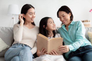 Küçük sevimli Asyalı kız, annesi ve büyükannesi ile birlikte evdeki kanepede kitap okuyor.