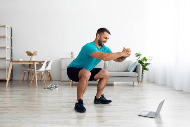 Mutlu Avrupalı adam bacak egzersizleri yapıyor, çömeliyor ve bilgisayar dersi izliyor.