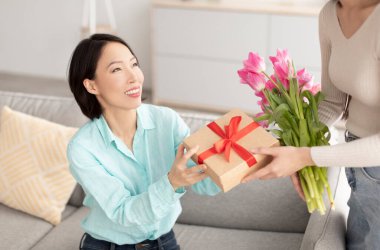 Genç bir kadın evinde olgun Asyalı annesine hediye paketi ve güzel çiçekler veriyor. Aile şenliği konsepti