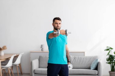 Ciddi milenyum Avrupalı erkek atlet mavi tişörtlü minimalist oturma odasında dambıl kaldırıyor