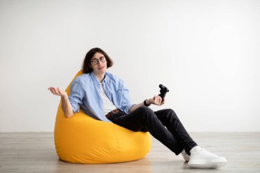 Sıkılmış adam elinde joystick ile puf minderinde oturuyor, video oyunları oynuyor ve kaybediyor, evde dinleniyor, boş yer var.