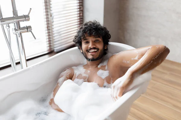 Bienestar, concepto de cuidado corporal. Feliz hombre indio relajado tomando baño de burbujas, mirando y sonriendo a la cámara, espacio libre — Foto de Stock