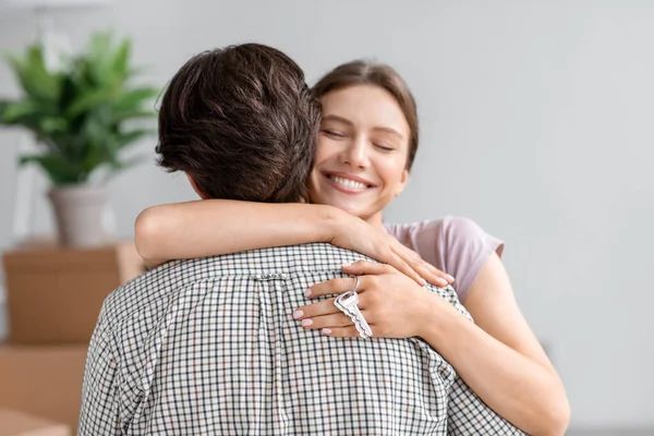Usmívající se šťastný pěkný mladý evropský manželka objímají manžela a ukazuje klíč z nového vlastního bytu v interiéru pokoje — Stock fotografie