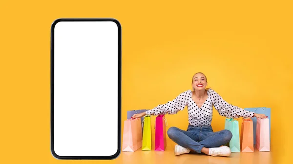 Mujer sentada cerca de grandes teléfonos inteligentes y bolsas de compras, fondo amarillo —  Fotos de Stock