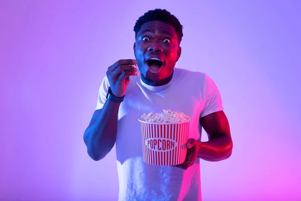 Portret podekscytowanego młodego czarnego faceta jedzącego popcorn z wiadra, oglądającego film w kinie lub w telewizji, neonowe światło — Zdjęcie stockowe