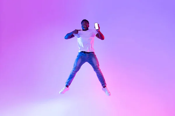 Pełna długość podekscytowany młody czarny facet skacze z telefonu komórkowego, wskazując na pusty ekran w świetle neonu, makieta — Zdjęcie stockowe
