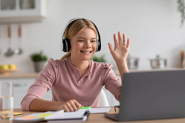 Heureux caucasien adolescent fille élève dans casque agitant main étudier avec ordinateur portable à table dans cuisine intérieur — Photo