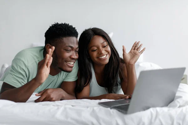 Alegre joven afroamericano esposa y marido yacen en la cama saludando la mano en la webcam ordenador en el dormitorio —  Fotos de Stock
