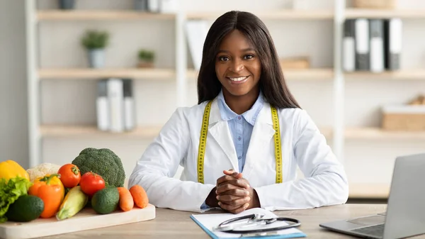 Porträt einer professionellen afrikanisch-amerikanischen Ernährungsberaterin, die am Arbeitsplatz in einer Gewichtsreduktionsklinik sitzt und in die Kamera lächelt — Stockfoto