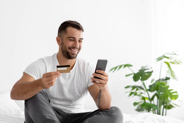 Lächelnder junger Mann in weißem T-Shirt kauft online ein, bestellt Waren per Smartphone und zeigt Kreditkarte — Stockfoto