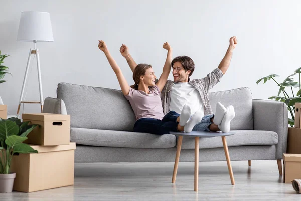 Счастливые взволнованные молодой муж и жена Европы радуются, поднимают руки на диван в гостиной интерьера — стоковое фото