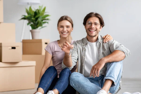 Tevreden blanke jongeman in bril en dame zitten op de vloer toon sleutels tussen kartonnen doos met spullen in de kamer — Stockfoto