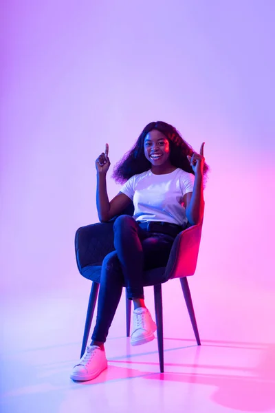 Sonriente joven mujer negra sentada en sillón, apuntando hacia arriba, ofreciendo espacio en blanco en luz de neón, maqueta para el anuncio — Foto de Stock