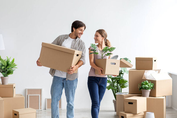 Laughing young caucasian couple carry cardboard boxes with stuff and plants in room interior