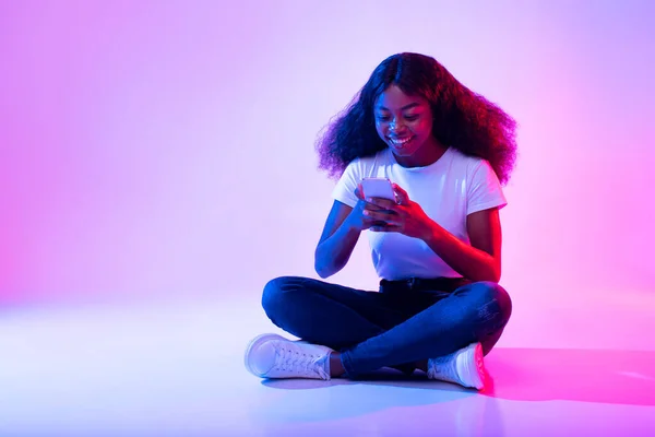 Joven mujer afroamericana sentada con las piernas cruzadas con el teléfono celular, navegar por las redes sociales, charlar con un amigo a la luz de neón — Foto de Stock