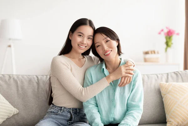 Retrato de la joven asiática y su madre madura abrazándose y sonriendo a la cámara, sentada en el sofá en casa — Foto de Stock