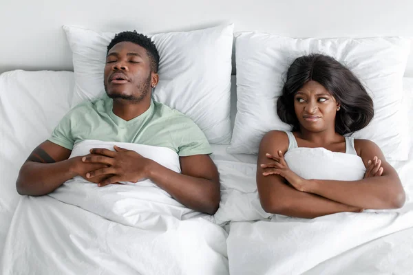 Незадоволена нещаслива афріканська жінка в Америці страждає від шуму і хропіння сплячого чоловіка на ліжку. — стокове фото