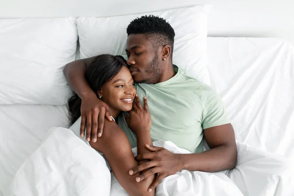 Alegre joven afroamericano marido abrazando esposa y besándose en la cama cómoda, descansando, relajarse en el dormitorio — Foto de Stock