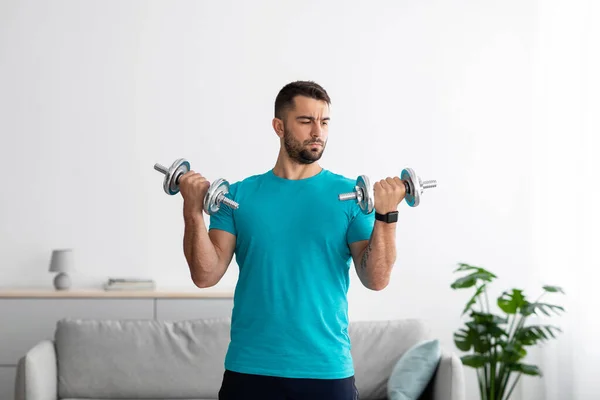 Серьезный спортсмен тысячелетия в синей футболке делает силовые упражнения для рук и поднимает гантели — стоковое фото