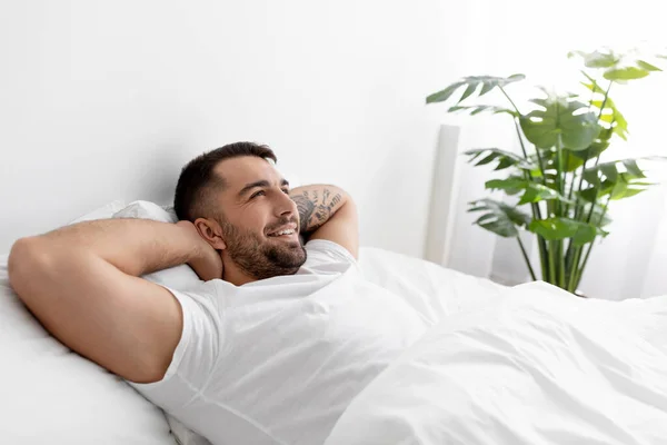 Happy Kaukasische jonge man in wit t-shirt rust en ontspant, kijkt naar lege ruimte op bed in slaapkamer interieur — Stockfoto