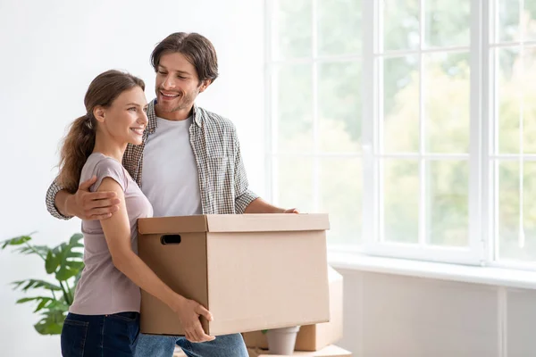 Sorrindo jovem marido europeu abraço esposa segurar caixa de papelão com coisas em quarto vazio interior — Fotografia de Stock