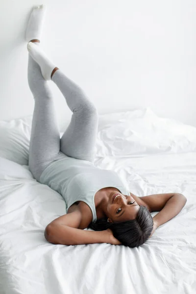 Весела афріканська жінка в домашньому одязі піднімає ноги на білу стіну і насолоджується вільним часом. — стокове фото