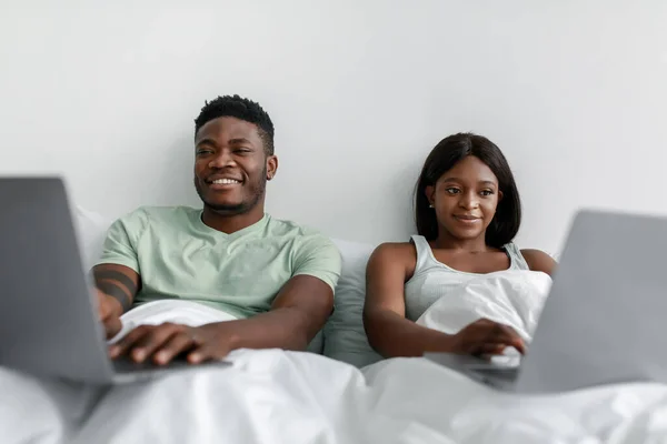 Ευτυχισμένο χιλιετίες αφροαμερικανός σύζυγος και σύζυγος ξαπλώνουν στο κρεβάτι με τους υπολογιστές, παίζουν παιχνίδια — Φωτογραφία Αρχείου