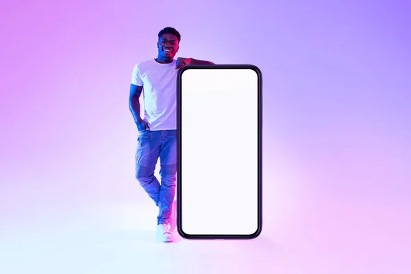 Gülümseyen genç siyahi adam cep telefonunun yanında duruyor. Boş beyaz ekranlı, neon ışıklı, mobil uygulamalı. — Stok fotoğraf
