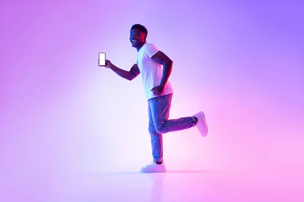 Pemuda kulit hitam yang bersemangat dengan smartphone berjalan dalam lampu neon, menunjukkan mockup untuk desain pada layar perangkat seluler — Stok Foto