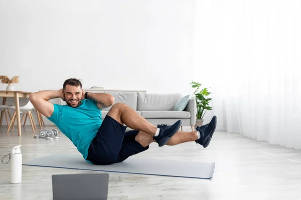 Веселый молодой европейский спортсмен в форме делает упражнения на прессе на коврике с ноутбуком в гостиной — стоковое фото