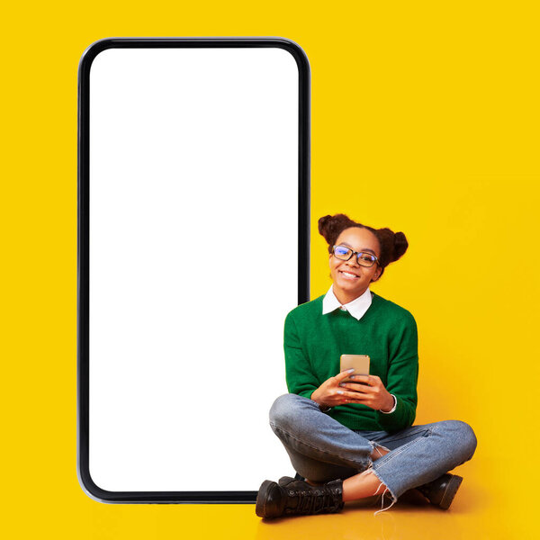 Африканская девочка-подросток, использующая телефон, сидящий рядом со смартфоном, желтый фон