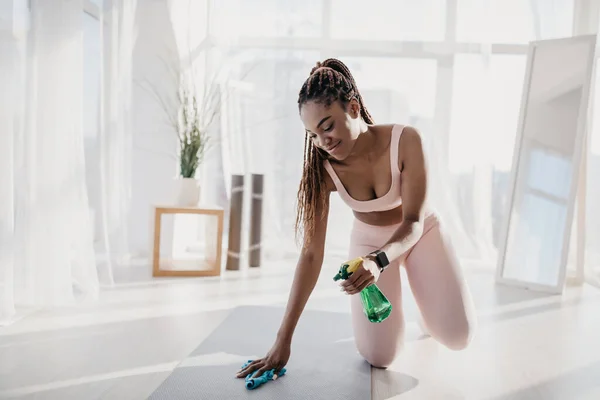 Desinfección de equipos deportivos. Mujer negra joven aplicando spray desinfectante y limpiando esterilla de yoga antes de entrenar en el gimnasio — Foto de Stock