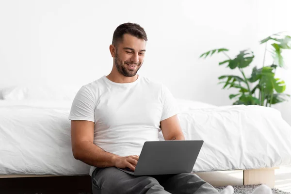 Wesoły atrakcyjne kaukaskie millenium mężczyzna do domu w białym t-shirt działa na laptopie siedzi obok łóżka — Zdjęcie stockowe