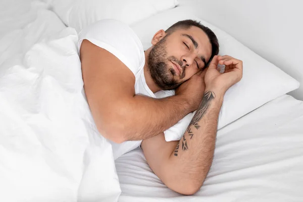 Hombre joven atractivo europeo en camiseta blanca duerme sobre una almohada suave en una cómoda cama en el interior del dormitorio —  Fotos de Stock