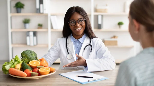 Дружелюбная черная женщина-диетолог консультирует пациента и демонстрирует тарелку со свежими фруктами и овощами — стоковое фото